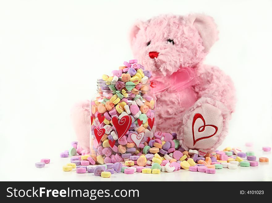 A soft, pink teddy bear sitting next to an overflowing container of conversation hearts. Shallow DoF with focus on the container of candy. Valentine concept with space for copy. A soft, pink teddy bear sitting next to an overflowing container of conversation hearts. Shallow DoF with focus on the container of candy. Valentine concept with space for copy.