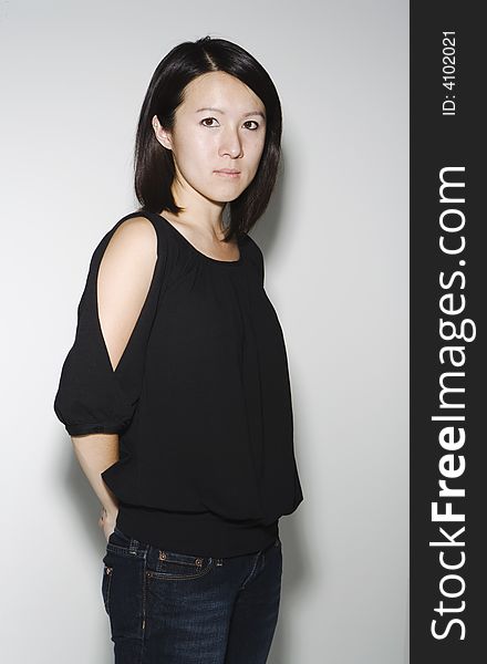 Asian woman against a white wall look directly into camera with her hand behind her back.  She is wearing a black top and denim jeans. Asian woman against a white wall look directly into camera with her hand behind her back.  She is wearing a black top and denim jeans.