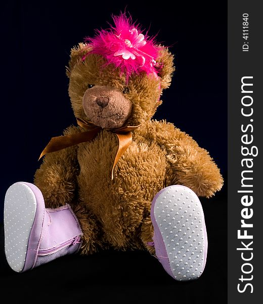 A brown teddybear with pink boots and pink feathers on its head. Black background.