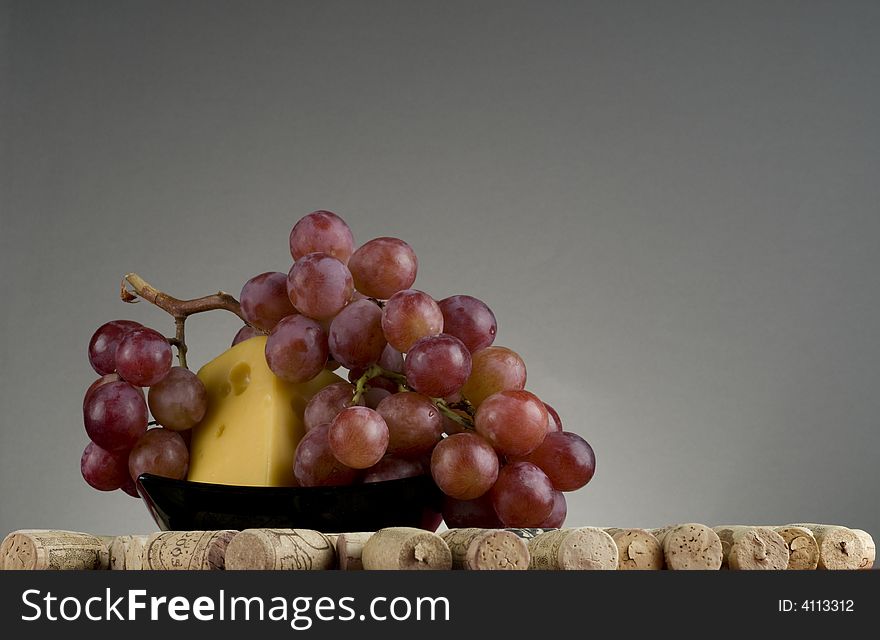 Wine cork with yellow cheese on black plate and red grape over grey background. Wine cork with yellow cheese on black plate and red grape over grey background.