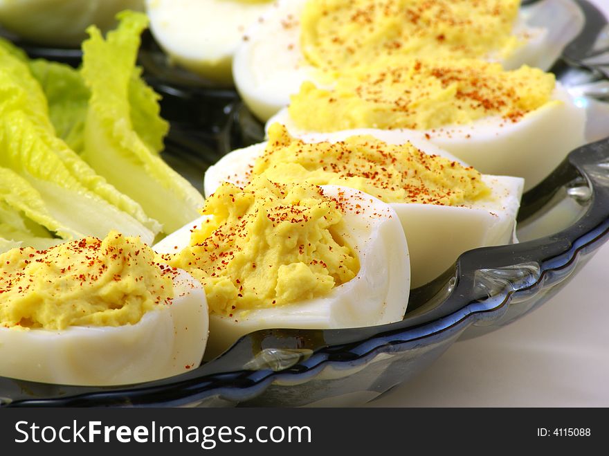 Homemade deviled eggs with a dash of cayenne pepper, displayed on depression era blue glass platter over white background. Small touch of baby lettuce completes the arrangement.