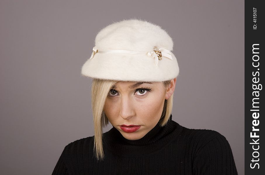 A blonde woman in a black turtleneck and a white fur hat pouting against a grey background. A blonde woman in a black turtleneck and a white fur hat pouting against a grey background