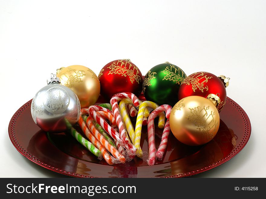 Shiny Christmas ornaments on red platter with wrapped candy canes in the middle. White background with copy space. Shiny Christmas ornaments on red platter with wrapped candy canes in the middle. White background with copy space.