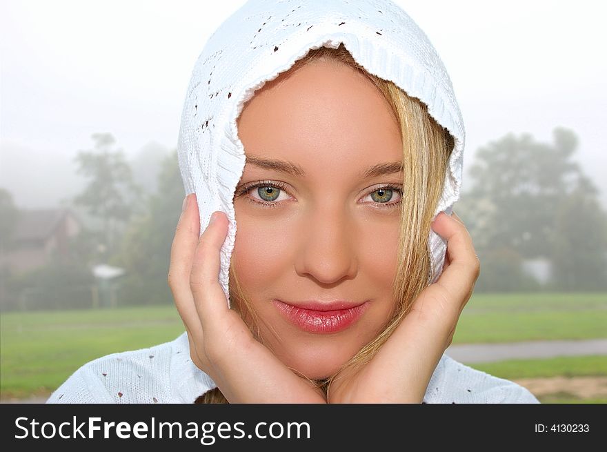 A beautiful young blonde woman wearing a knitted white hood, with her hands clasping the sides of her head while holding a friendly expression. Foggy park in the background. A beautiful young blonde woman wearing a knitted white hood, with her hands clasping the sides of her head while holding a friendly expression. Foggy park in the background.