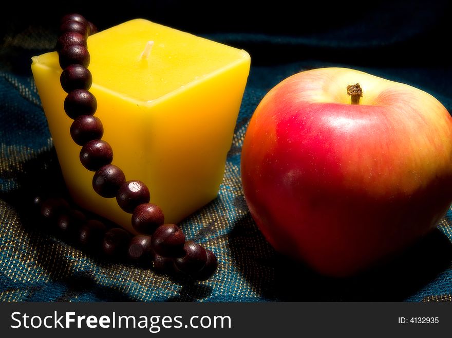 Red apple yellow candle and wooden necklace on textile background macro studio. Red apple yellow candle and wooden necklace on textile background macro studio