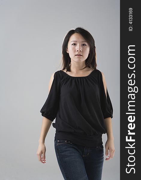 Young Asian woman wearing a black top and denim jeans walks forward with her eyes looking at the viewer against a gray studio background. Young Asian woman wearing a black top and denim jeans walks forward with her eyes looking at the viewer against a gray studio background.