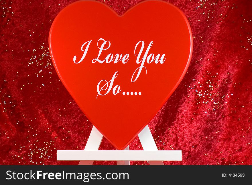 A large red heart on a wooden easel or stand with the message, I love you so... against a red velvet background with sequins. A large red heart on a wooden easel or stand with the message, I love you so... against a red velvet background with sequins.