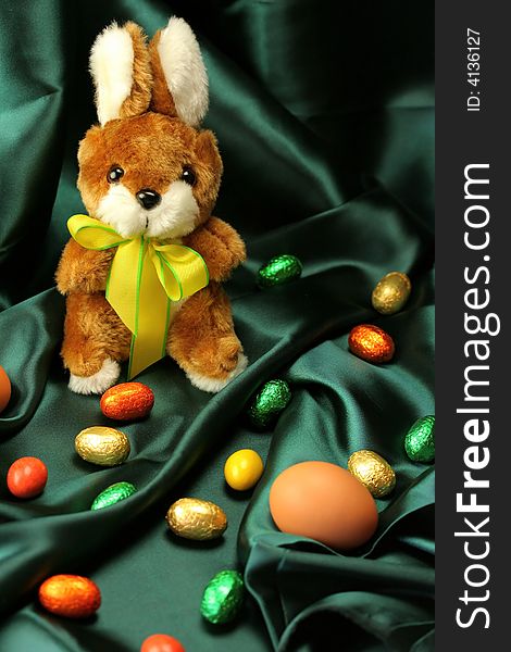 Easter theme - a fluffy bunny (rabbit) with a yellow ribbon sitting on a dark green satin fabric - textile, surrounded by colorful Easter eggs. Easter theme - a fluffy bunny (rabbit) with a yellow ribbon sitting on a dark green satin fabric - textile, surrounded by colorful Easter eggs.