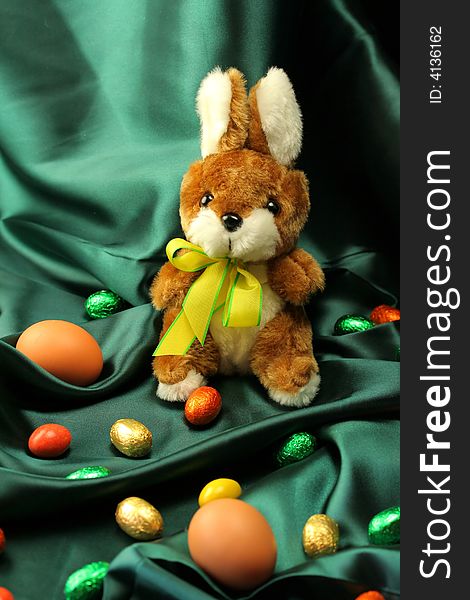 Easter theme - a fluffy bunny (rabbit) with a yellow ribbon sitting on a dark green satin fabric - textile, surrounded by colorful Easter eggs. Easter theme - a fluffy bunny (rabbit) with a yellow ribbon sitting on a dark green satin fabric - textile, surrounded by colorful Easter eggs.