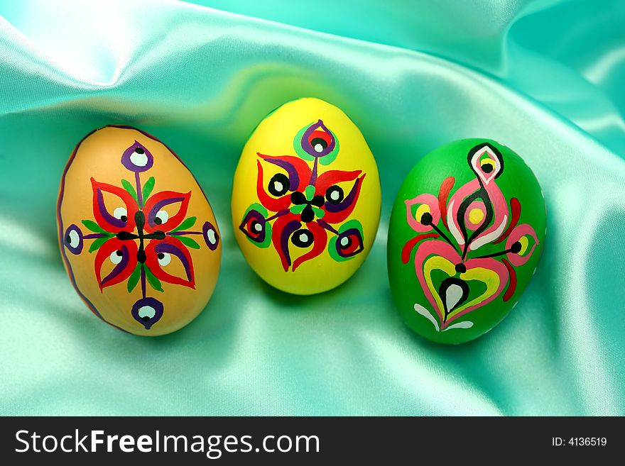 Three handpainted Easter eggs (natural, yellow and green) lying on turquoise satin fabric - background. Three handpainted Easter eggs (natural, yellow and green) lying on turquoise satin fabric - background.