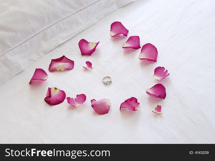 A pink heart made of rose petals on a crisp white bed enclose a diamond engagment ring. A pink heart made of rose petals on a crisp white bed enclose a diamond engagment ring