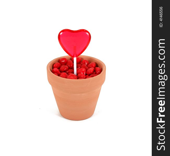 A red, heart shaped lollipop grows out of cinnamon heart soil in a terracotta flowerpot. Isolated on white background. A red, heart shaped lollipop grows out of cinnamon heart soil in a terracotta flowerpot. Isolated on white background.