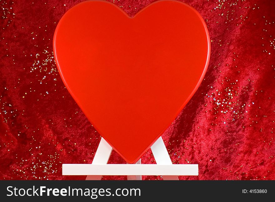 A large red heart on a wooden easel or stand with large open, blank space for copy or text.  Red background with sparkling sequins. A large red heart on a wooden easel or stand with large open, blank space for copy or text.  Red background with sparkling sequins.