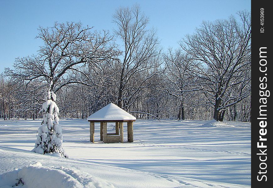 A gazebo with a water fountain inside covered with snow surrounded by trees. A gazebo with a water fountain inside covered with snow surrounded by trees.