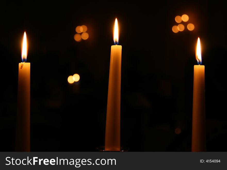 Three candles burning at night with other lights out of focus in the background