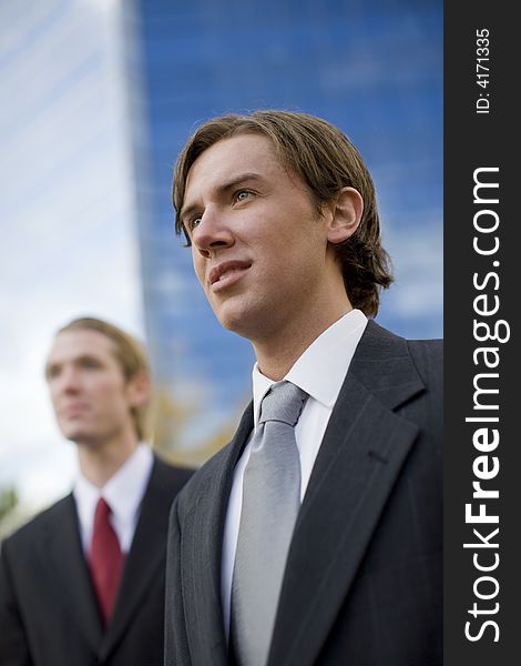 Two businessmen standing side by side looking forward in front of tall blue office building. Two businessmen standing side by side looking forward in front of tall blue office building