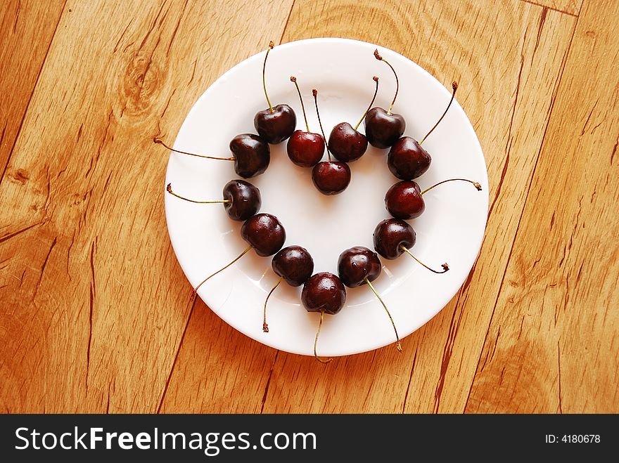 Red cherries in heart shape on the white plate, standing on the wooden table. Red cherries in heart shape on the white plate, standing on the wooden table