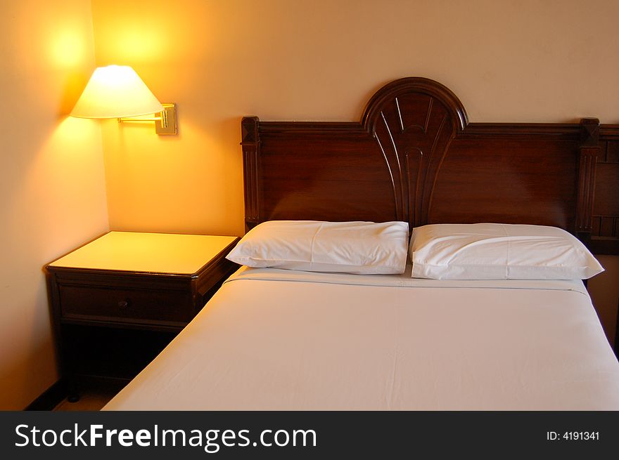 Hotel bedroom interior containing bed covered with white bedsheet, pillows and bed lamp.