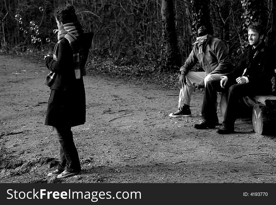 Three friends in a park: the two men are seated and the young lady is making soap bubbles. (black and white). Three friends in a park: the two men are seated and the young lady is making soap bubbles. (black and white)