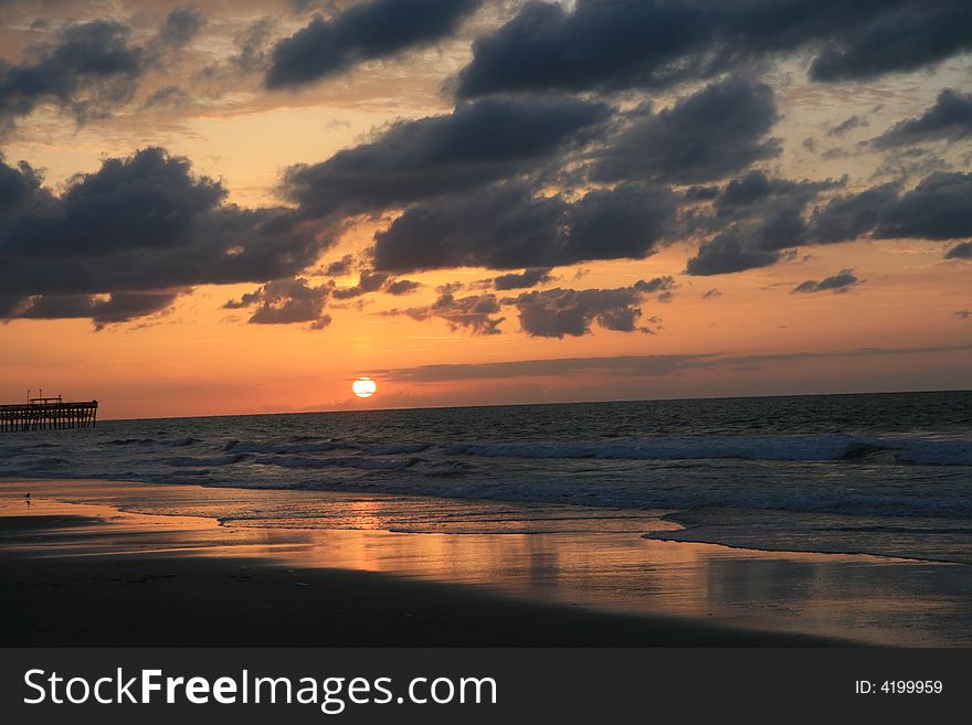 Early morning summer sunrise over ocean with sun coming up. Clouds are low and dark and sea is calm. A pier is visible in the distance at the mid-bottom left of the frame. Early morning summer sunrise over ocean with sun coming up. Clouds are low and dark and sea is calm. A pier is visible in the distance at the mid-bottom left of the frame.