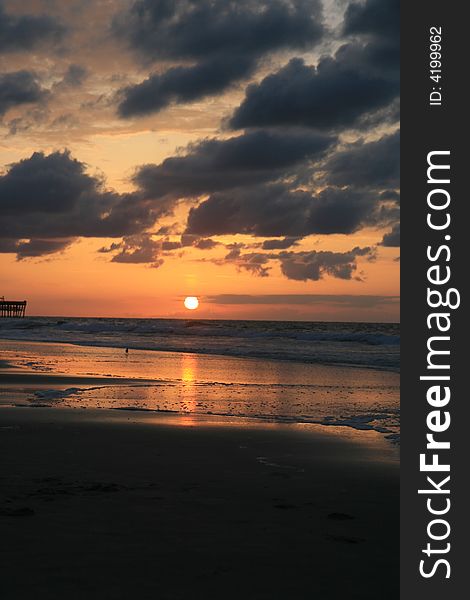 Early morning summer sunrise over ocean with sun about coming up. Clouds are low and dark and sea is calm. A pier is visible in the distance at the middle left of the frame. Early morning summer sunrise over ocean with sun about coming up. Clouds are low and dark and sea is calm. A pier is visible in the distance at the middle left of the frame.