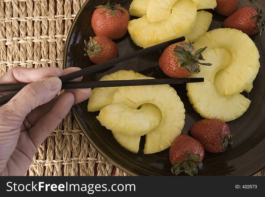 Close up of juicy yellow pineapple rings and red, plump strawberries and hand using chopsticks to pick up the strawberry. Close up of juicy yellow pineapple rings and red, plump strawberries and hand using chopsticks to pick up the strawberry.