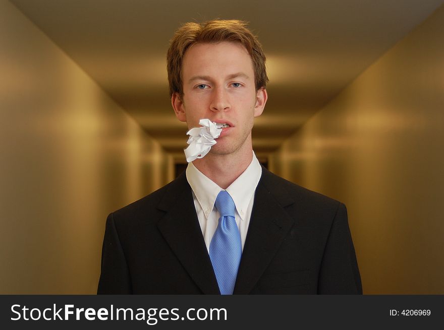 A man in a black suit stands in an empty hallway with a crumpled piece of paper in his mouth. A man in a black suit stands in an empty hallway with a crumpled piece of paper in his mouth