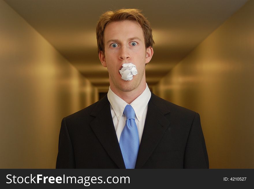 A man in a black suit stands in an empty hallway with a crumpled piece of paper in his mouth looking surprised. A man in a black suit stands in an empty hallway with a crumpled piece of paper in his mouth looking surprised