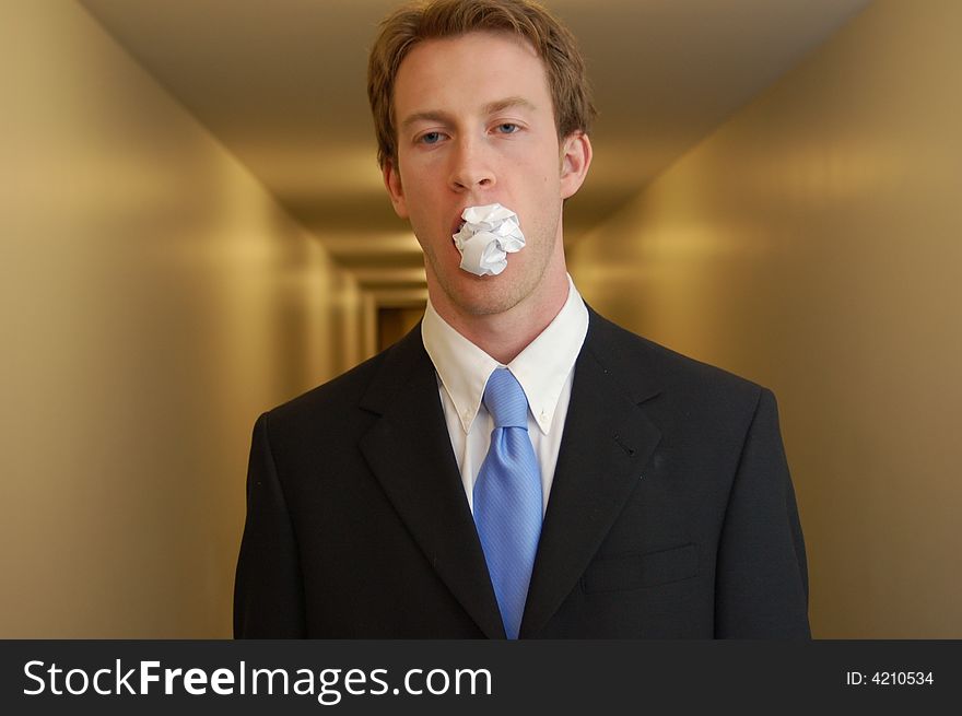 A man in a black suit stands in an empty hallway with a crumpled piece of paper in his mouth looking surprised. A man in a black suit stands in an empty hallway with a crumpled piece of paper in his mouth looking surprised