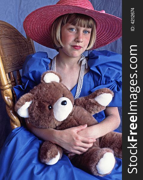 Young girl with bright blue eyes playing dress up in blue prom gown with big brimmed pink straw hat. Sitting in chair holding her brown and white teddy bear. Looking a bit forlorn. Young girl with bright blue eyes playing dress up in blue prom gown with big brimmed pink straw hat. Sitting in chair holding her brown and white teddy bear. Looking a bit forlorn.