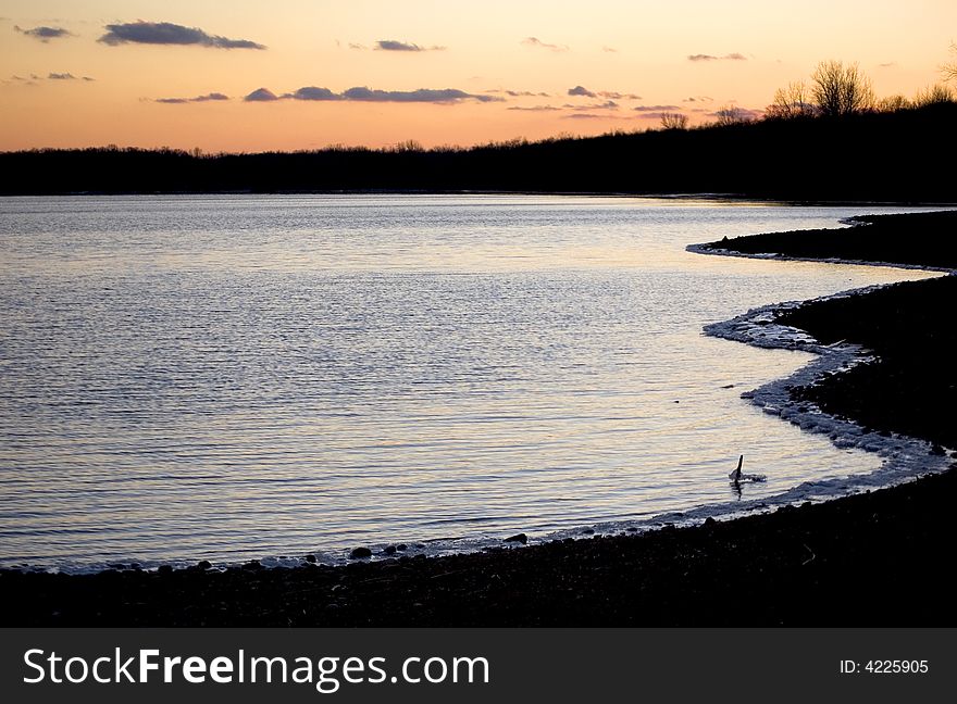 A tranquil scene of a winter sunset over a calm lake, partially iced at the water's edge. A tranquil scene of a winter sunset over a calm lake, partially iced at the water's edge.