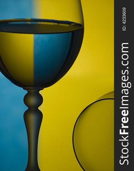 Glass of water on the background divided by yellow and blue colour together with a torso of recumbent glass. Glass of water on the background divided by yellow and blue colour together with a torso of recumbent glass