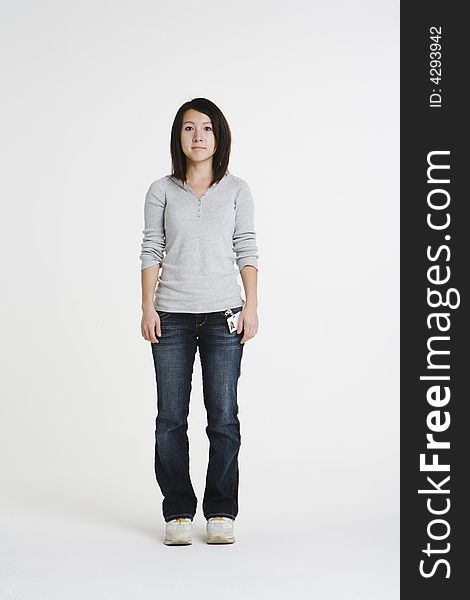 Young Asian woman with a office badge stands in a white studio wearing casual attire. She is wearing a gray shirt and denim jeans with a blank look on her face. Young Asian woman with a office badge stands in a white studio wearing casual attire. She is wearing a gray shirt and denim jeans with a blank look on her face.