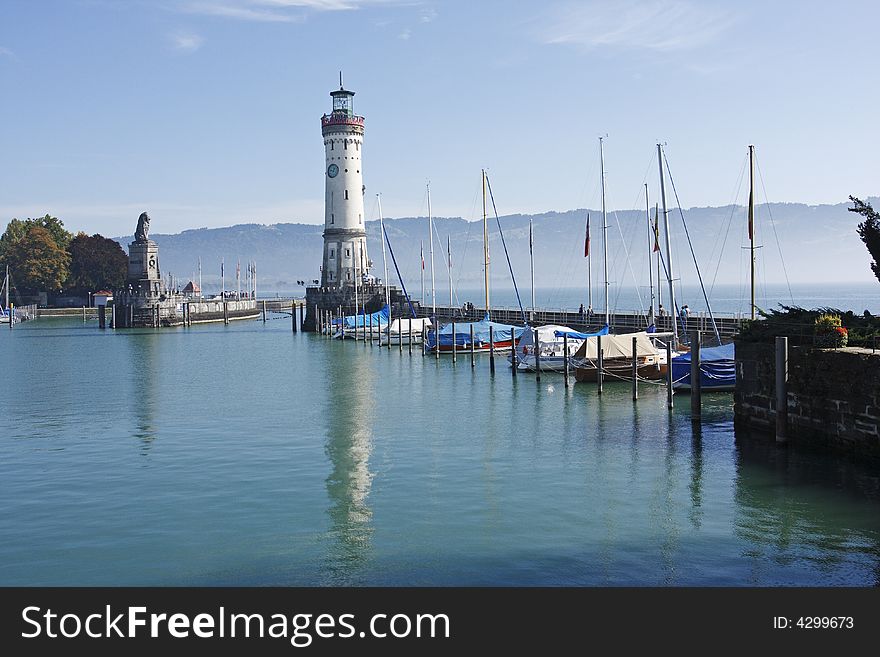 Lindau harbour looking at the harbour entrance guarded by a lighthouse and statue of a lion. Sailing boats are moored along the breakwater and the lighthouse is reflected in the calm water. Lake Constance is covered in early morning mist. Lindau harbour looking at the harbour entrance guarded by a lighthouse and statue of a lion. Sailing boats are moored along the breakwater and the lighthouse is reflected in the calm water. Lake Constance is covered in early morning mist.