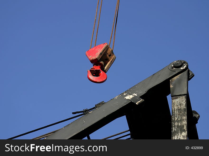 Red lifting hook suspended onfour twisted streel cables hanging over a black steel crane boom frame. Lots of blue sky for text. Red lifting hook suspended onfour twisted streel cables hanging over a black steel crane boom frame. Lots of blue sky for text.