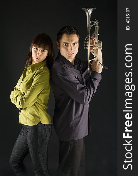 The woman and the man standing back to each other's. The man holding trumpet in hands. They're looking at camera. Side view. The woman and the man standing back to each other's. The man holding trumpet in hands. They're looking at camera. Side view.