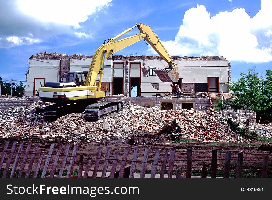 Yellow bulldozer turning old building into rubble beneath a brilliant summer sky. Huge debris pile and construction fence in foreground. Yellow bulldozer turning old building into rubble beneath a brilliant summer sky. Huge debris pile and construction fence in foreground.