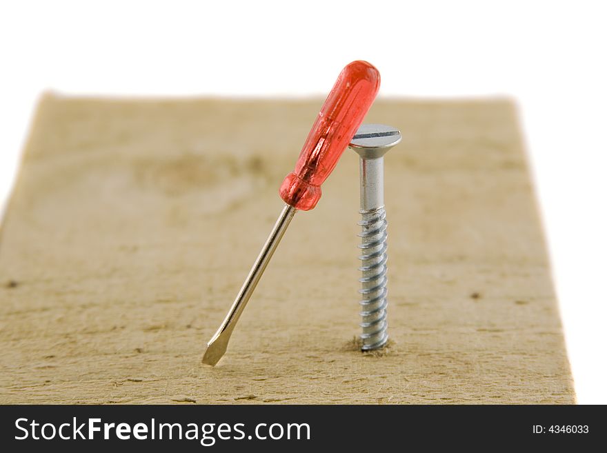 A large screw, screwed into a plank of wood with a very small, red-handled screwdriver leaning against it.