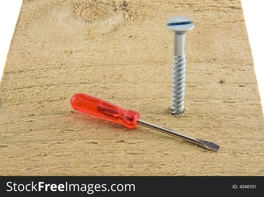 A very small, red-handled screwdriver liying on a plank of wood in which is a large screw.