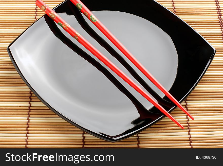 Red chopsticks and black dish on a bamboo mat. Close-up. Variant one.