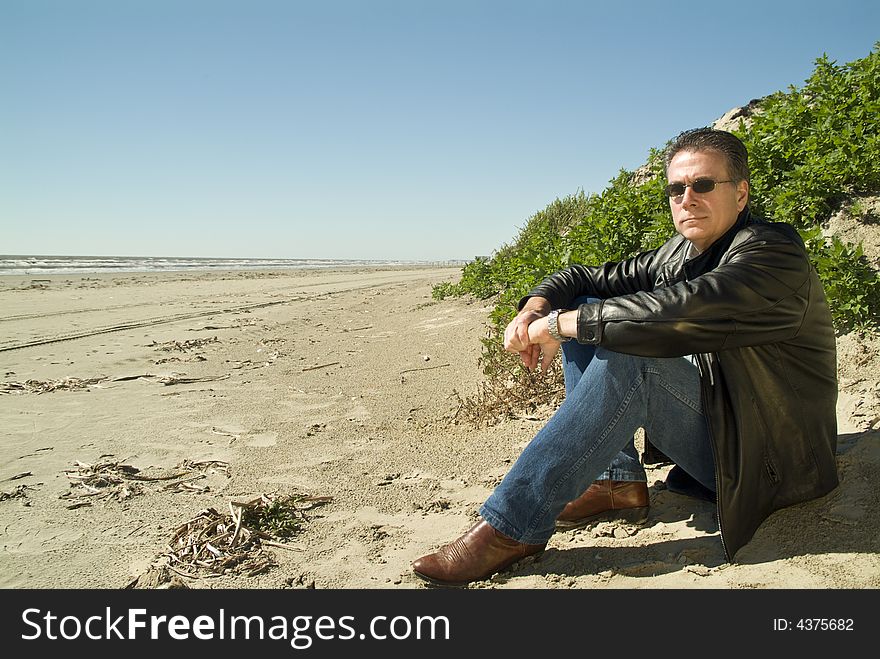 A man sitting at the edge of a sand dune gazing out at the beach in front of him. A man sitting at the edge of a sand dune gazing out at the beach in front of him.