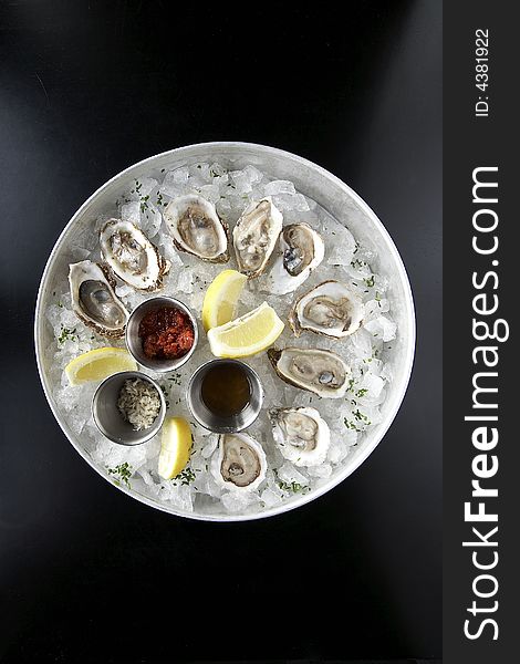 A seafood appetizer of clams on a black table shot from above. The clams are in a tray of ice with lemons and sauce.