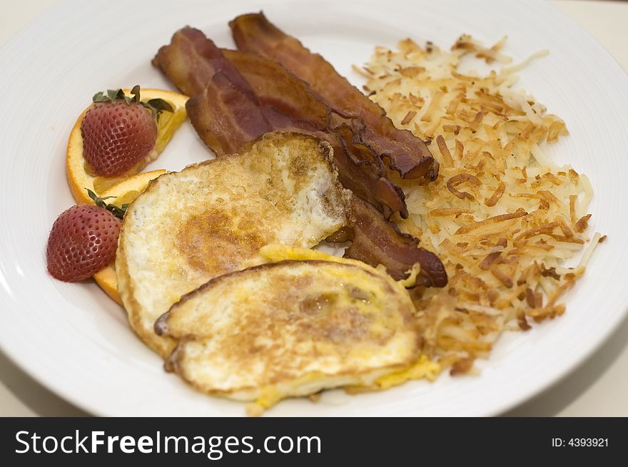 Bacon and fried egg breakfast with hash browns and fruit.  Shallow depth of field.