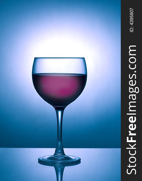 Image of a glass of dark wine silhouetted against a blue spotlight on dark background with reflection. Image of a glass of dark wine silhouetted against a blue spotlight on dark background with reflection
