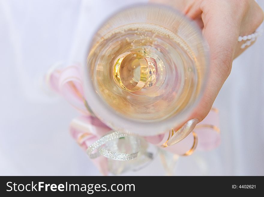 A hand of a bride holds a glass full of champagne and two gold rings in it. A hand of a bride holds a glass full of champagne and two gold rings in it