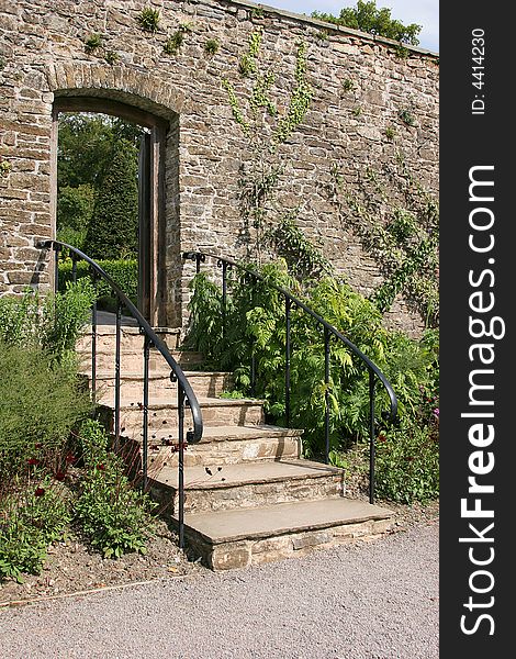Ancient garden stairway with metal handrails, set within an old stone wall and leading to a garden beyond. Flowers and shrubs either side of the steps.