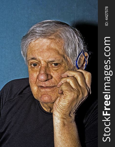 Dramatic side portrait of a senior man with a pair of glasses in his hand next to his face wearing a black shirt isolated on gray/blue. Dramatic side portrait of a senior man with a pair of glasses in his hand next to his face wearing a black shirt isolated on gray/blue