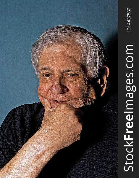 Dramatic portrait of a senior man with his hand on the side of his face wearing a black shirt isolated on gray/blue