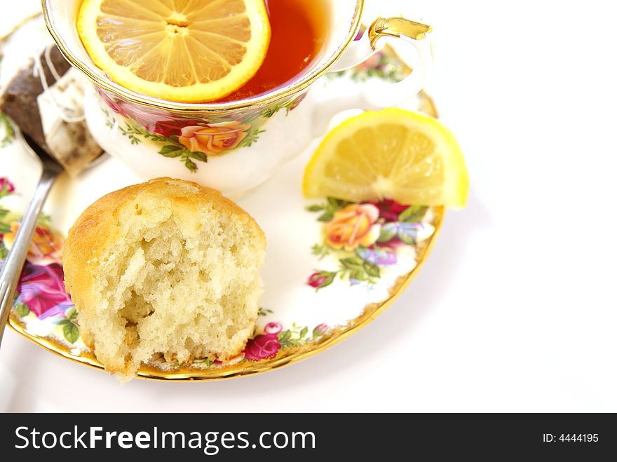 Partially eaten cake muffin on saucer next to tea, lemon, and spoon.