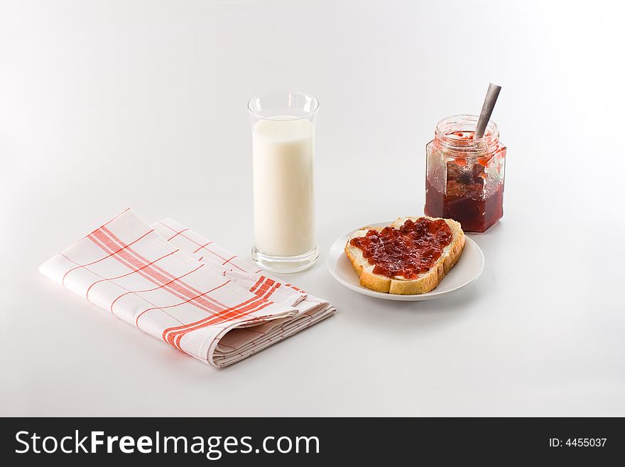 Breakfast scene: glass of milk, a slice of bread with butter and jam on a small plate, a jam jar with a teaspoon and a red striped kitchen cotton towel isolated on white background. Breakfast scene: glass of milk, a slice of bread with butter and jam on a small plate, a jam jar with a teaspoon and a red striped kitchen cotton towel isolated on white background.